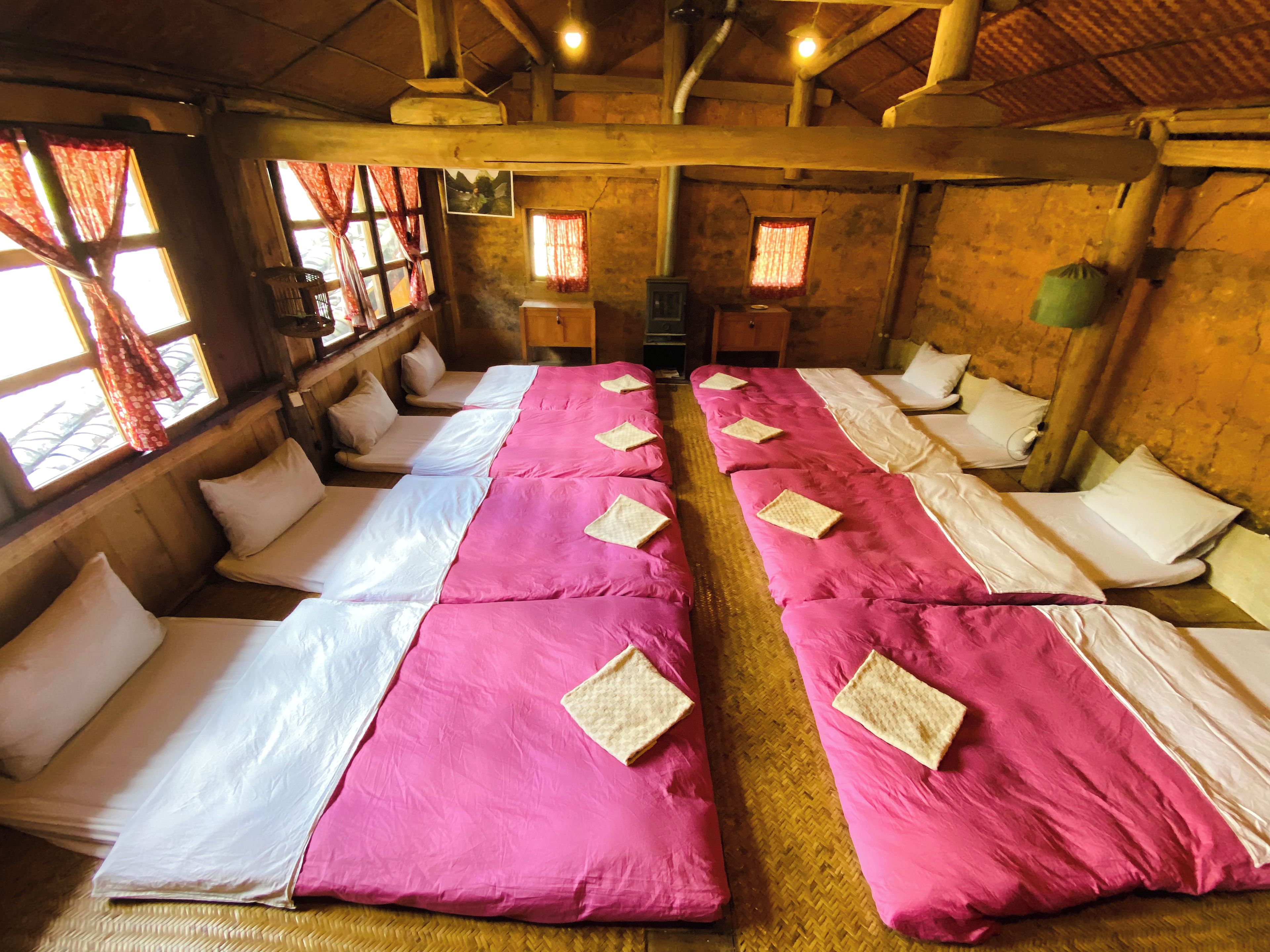 Ein Zimmer mit mehreren in Reihen angeordneten Betten, Holzbalken an der Decke und fensterbepflanzung mit Vorhängen.