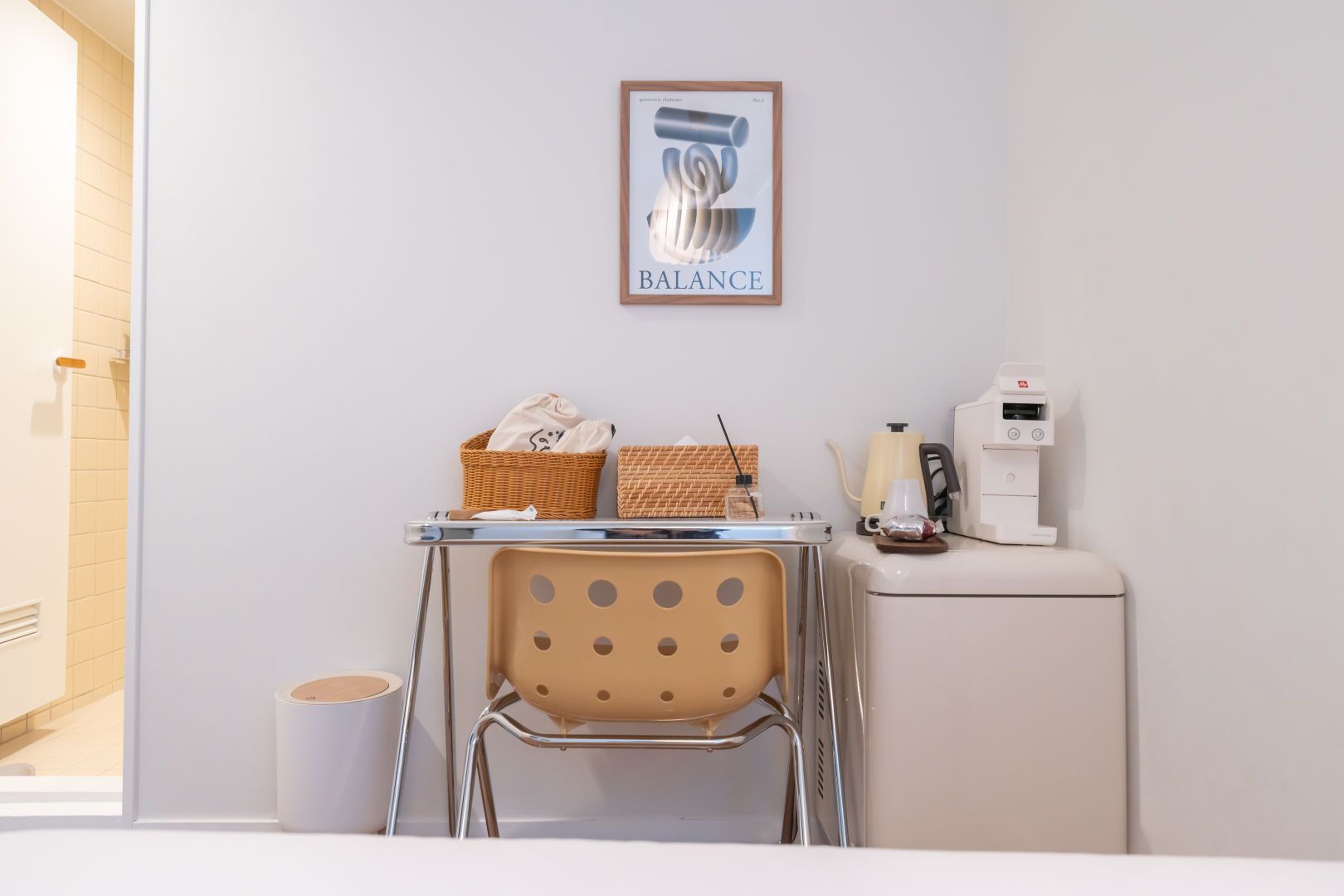 Quarto limpo e moderno, com parede branca, uma obra de arte emoldurada intitulada 'BALANCE', uma pequena mesa com uma cadeira, uma máquina de café e uma lixeira.