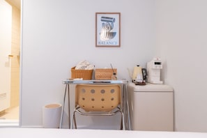 Una habitación limpia y moderna con pared blanca, una obra enmarcada titulada 'BALANCE', una mesita con una silla, una cafetera y un cubo de basura.