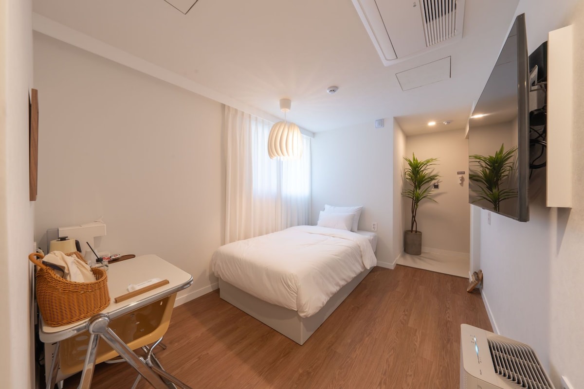 A hotel room with a bed, a desk, a chair, a plant, and a TV mounted on the wall.