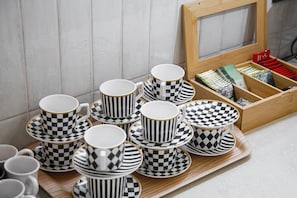 Una bandeja con varias tazas y platillos blancos y negros a cuadros, una caja de madera con pinceles y un paquete rojo, y una pared de azulejos al fondo.