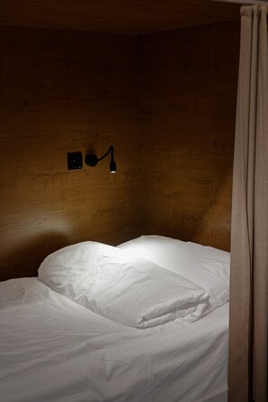 Una cama individual con ropa de cama blanca en una habitación con paredes de madera y una lámpara fijada a la pared.