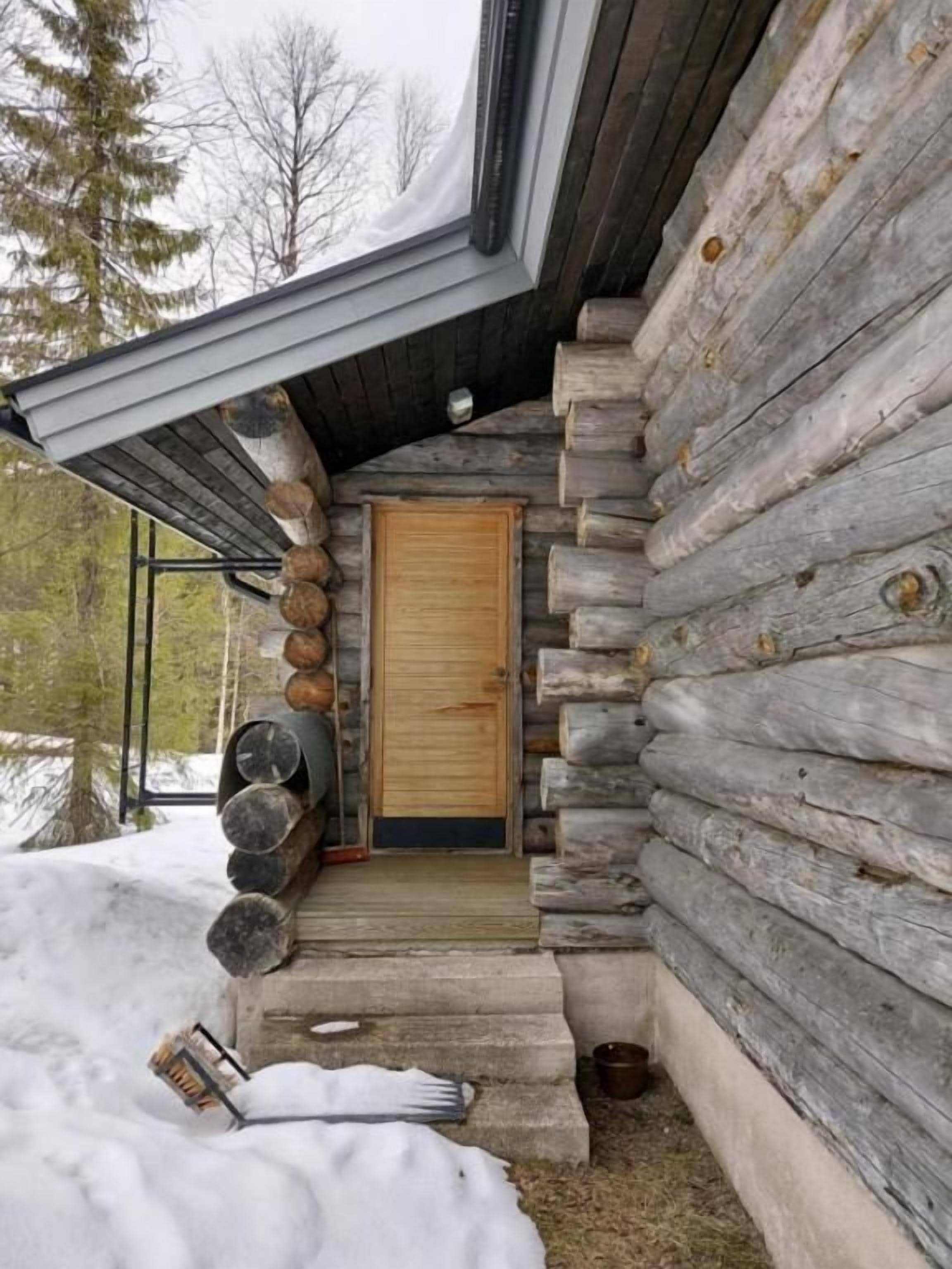 Een blokhut met een houten deur, een veranda met houtconstructies en een met sneeuw bedekte grond.