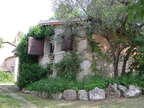Edifici de pedra rústic amb una porta i persianes de fusta, envoltat de vegetació i un mur de pedra.