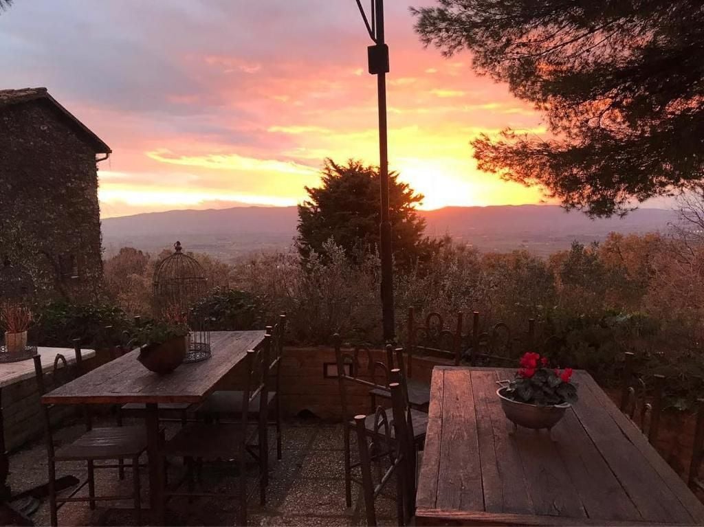 A rustic outdoor dining area with wooden tables and chairs, surrounded by vegetation and a view of distant hills.