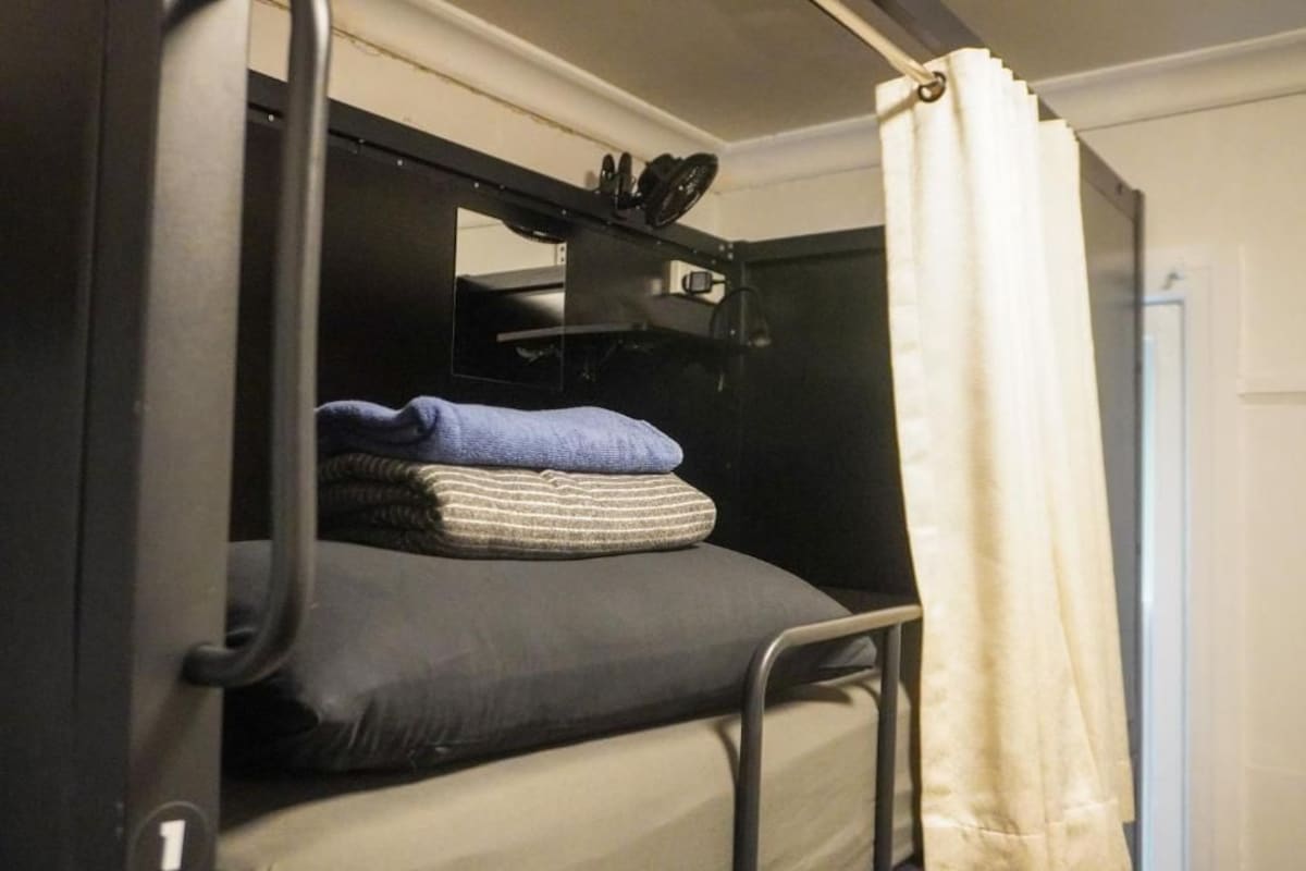 Een stapelbed met hoofdeinde, een klein plankje met spullen, en een gedeeltelijk dichtgetrokken gordijn.