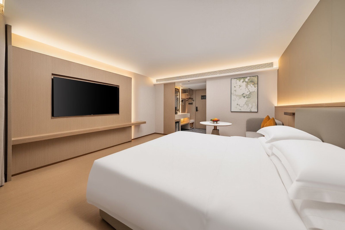 Una camera d'albergo con un letto grande, una TV a schermo piatto fissata al muro, un tavolino rotondo con una ciotola di frutta e uno specchio.