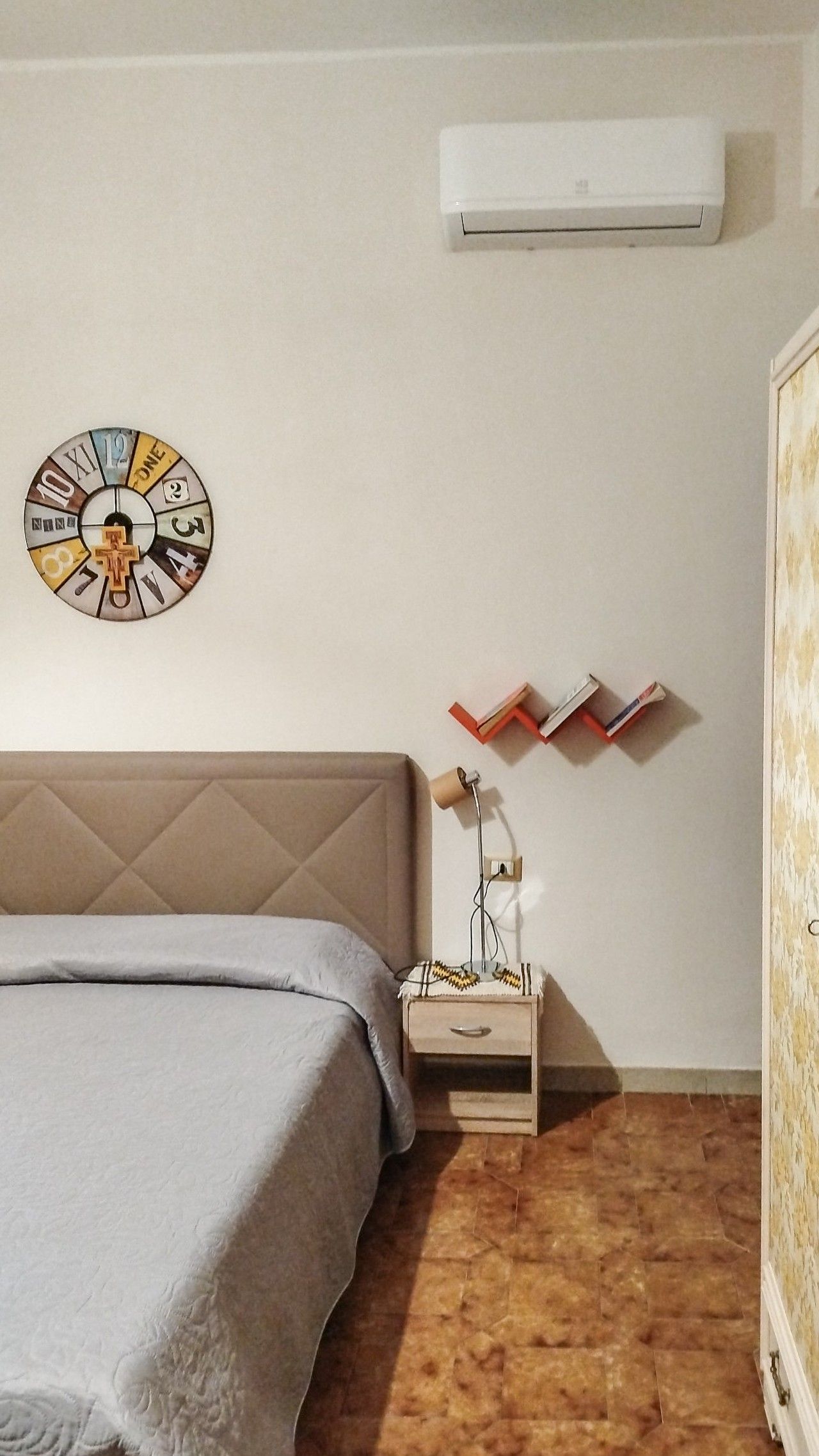 Ein Schlafzimmer mit einem Bett, einer Wanduhr, einem Nachttisch mit Büchern und einer Klimaanlage.