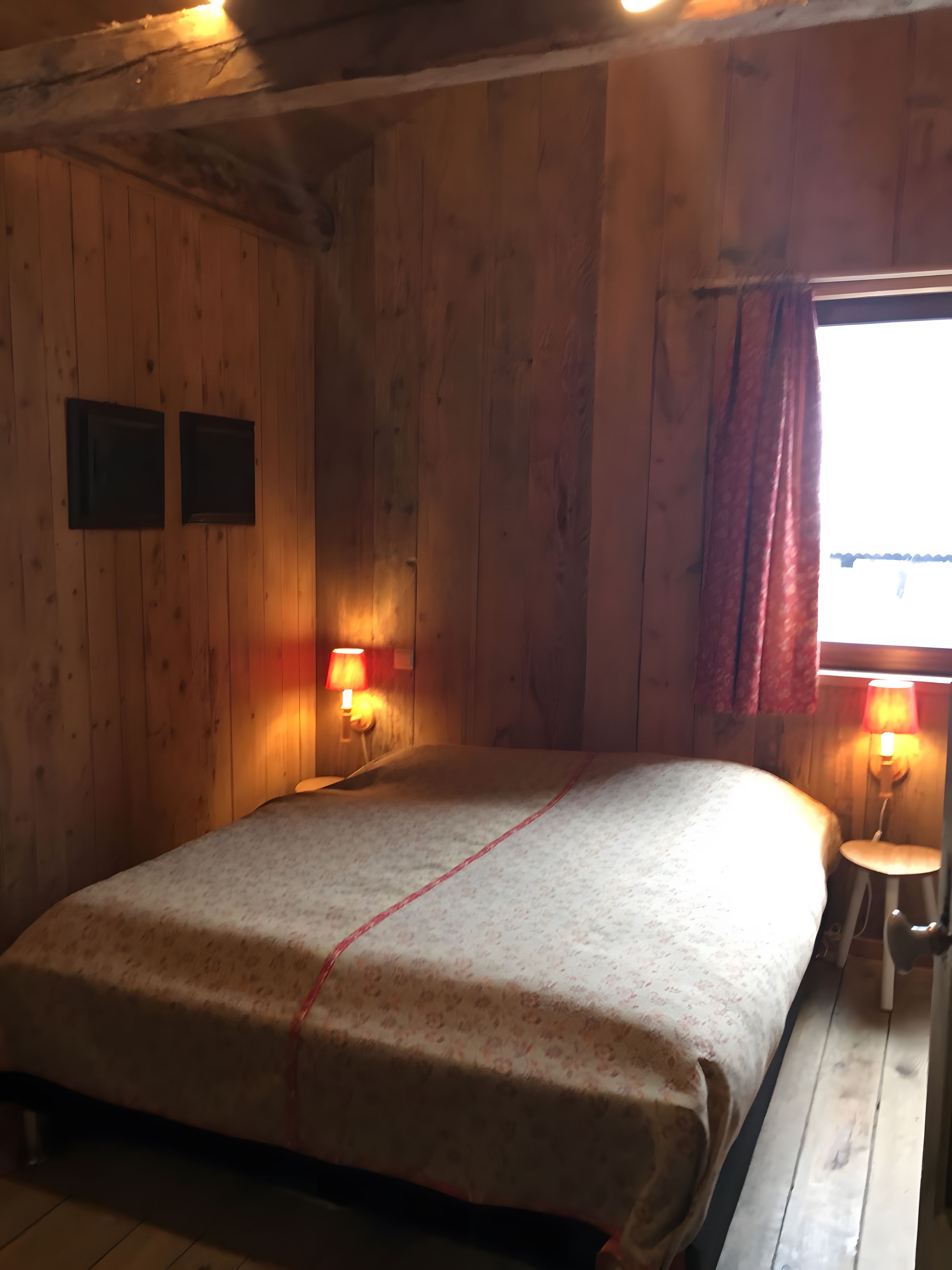 Une pièce en bois avec un lit, deux lampes fixées au mur et une fenêtre avec des rideaux.