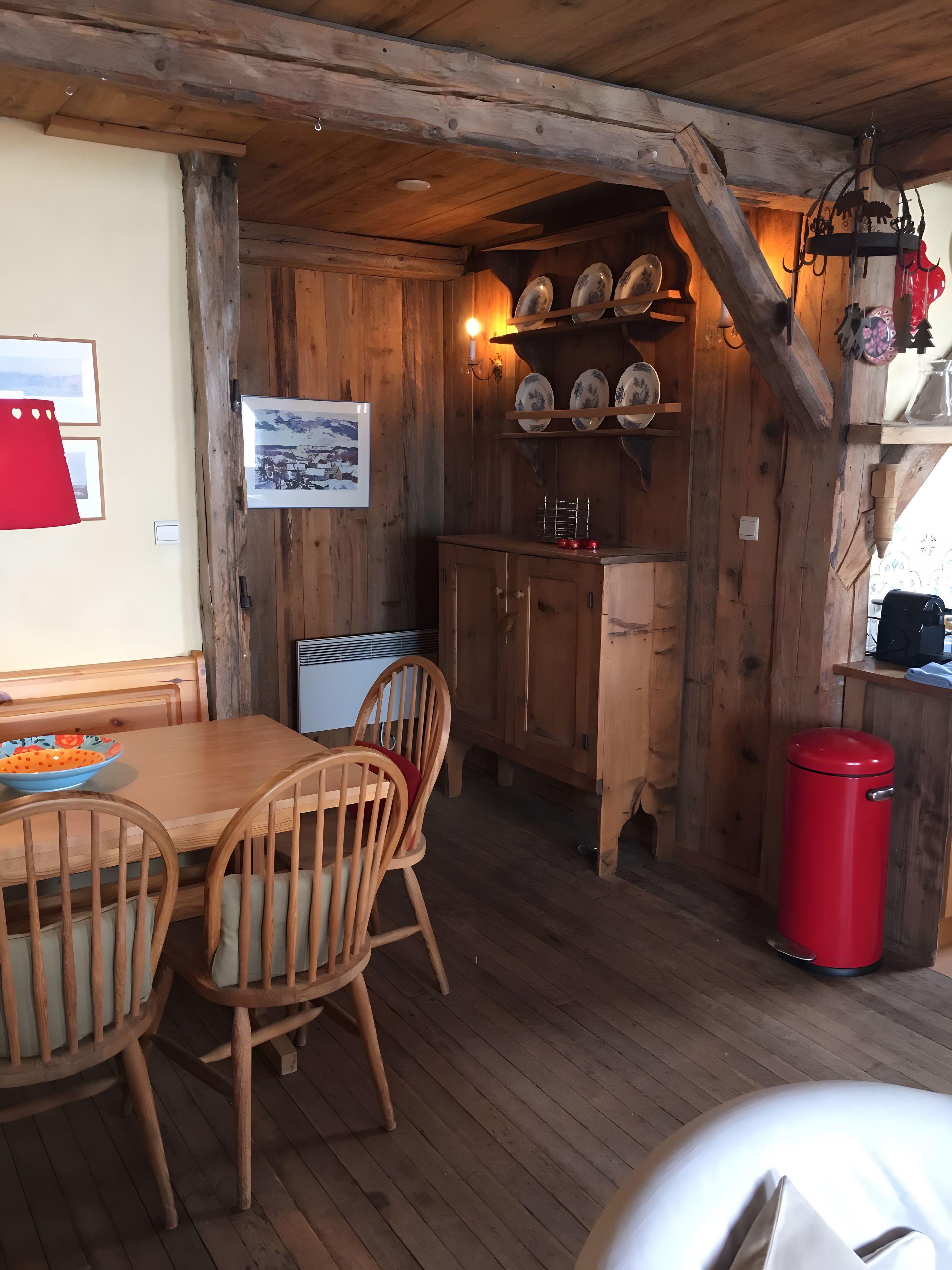 A wooden interior with a dining table, chairs, a red trash bin, and a wall-mounted shelf with plates.