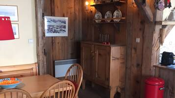 A wooden interior with a dining table, chairs, a red trash bin, and a wall-mounted shelf with plates.