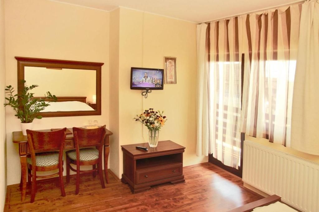 A room with a wooden desk, two chairs, a television mounted on the wall, a vase with flowers on a small table, and a window with curtains.