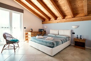 A bedroom with a wooden ceiling, a bed with patterned bedding, a wicker chair, a bedside table, and a nightstand.