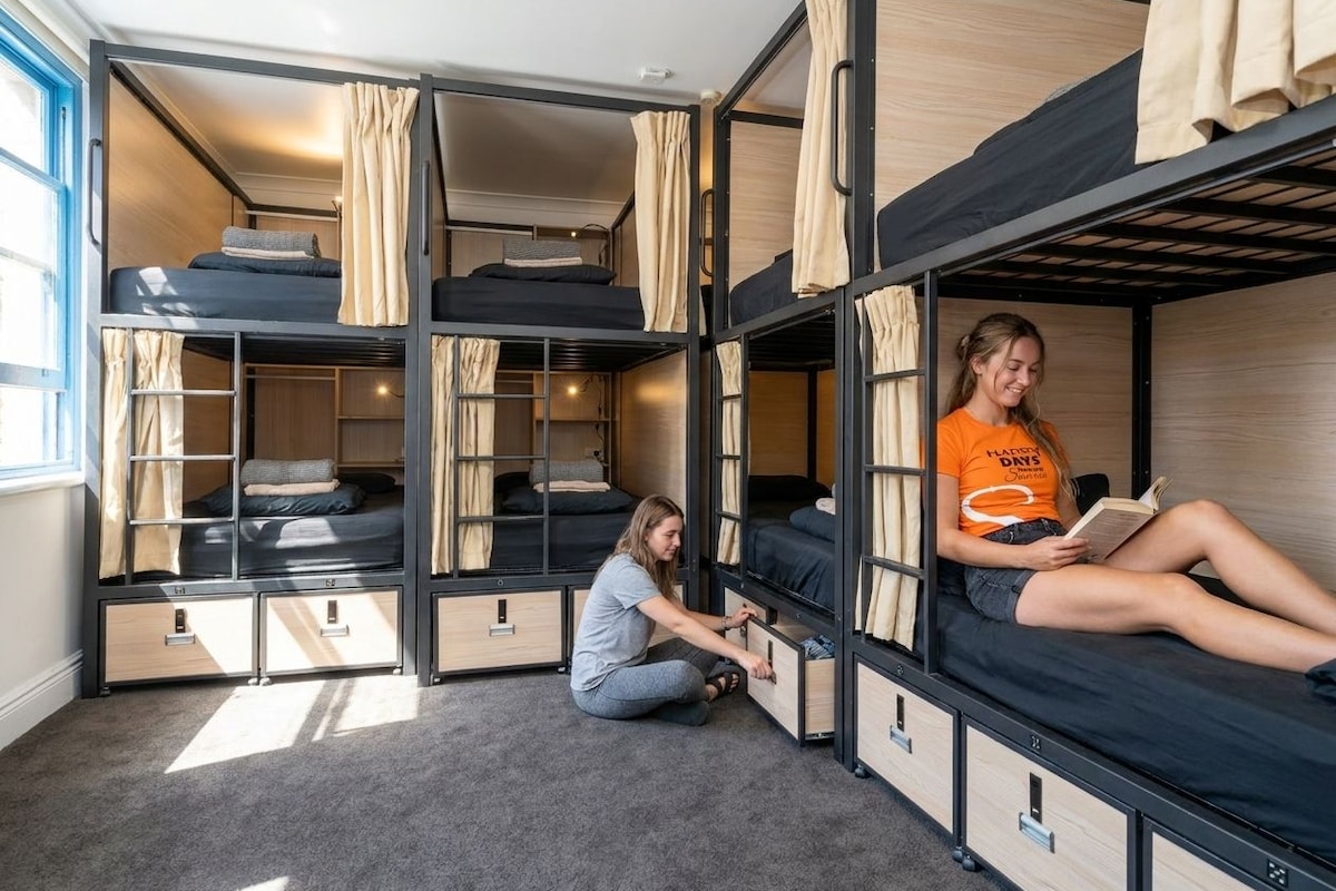Une chambre avec un lit superposé, une fenêtre, une personne qui lit et une autre assise sur le sol.