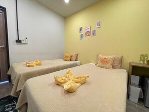 A hotel room with two beds, each with a towel folded in a bow shape. There are wall-mounted picture frames and a small table with a lamp and a bottle. - Good Sleep House (Ban Ta Khun)