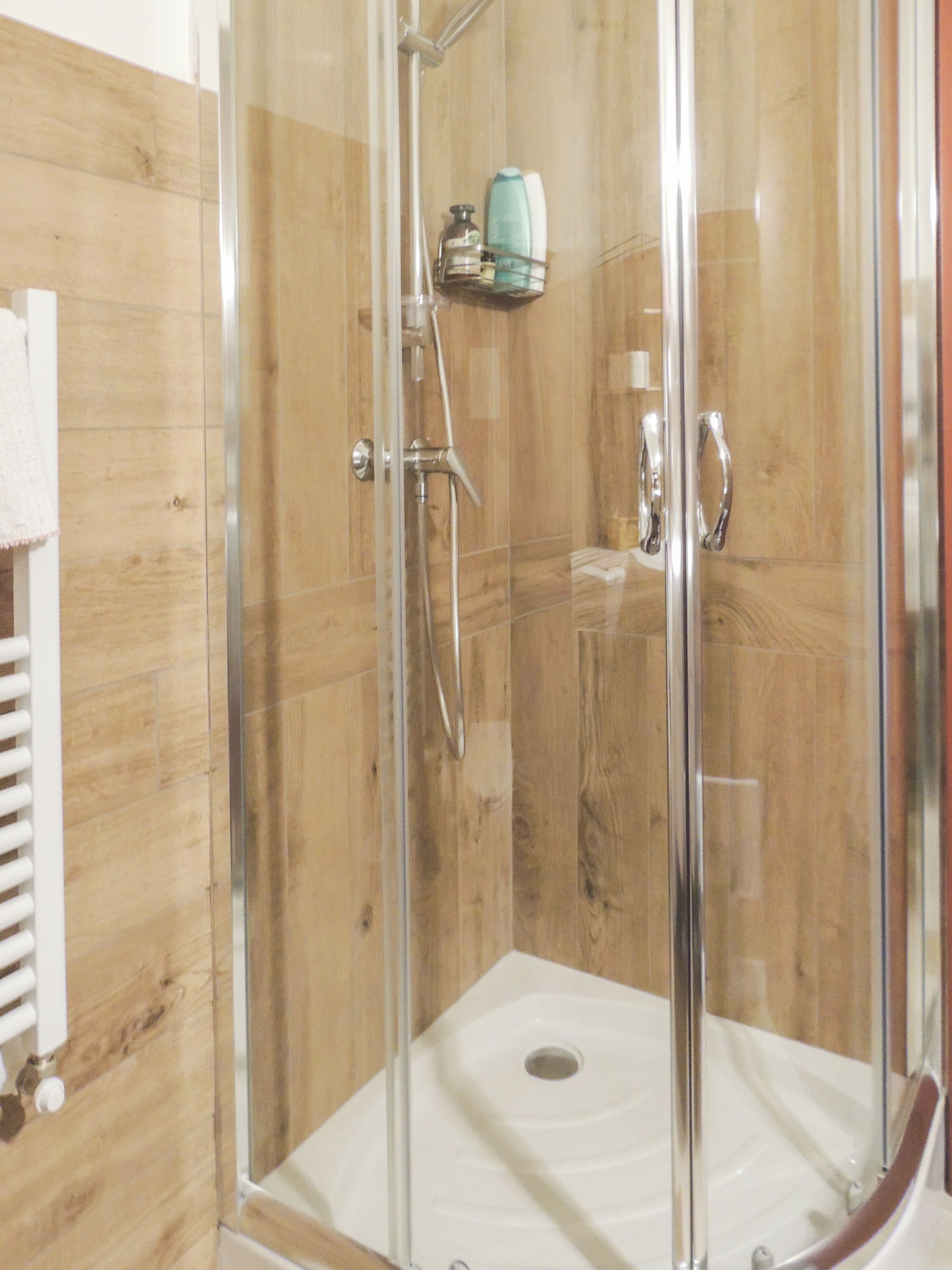 A glass-enclosed shower stall with a wooden wall backdrop, a white sink, and a towel rack.