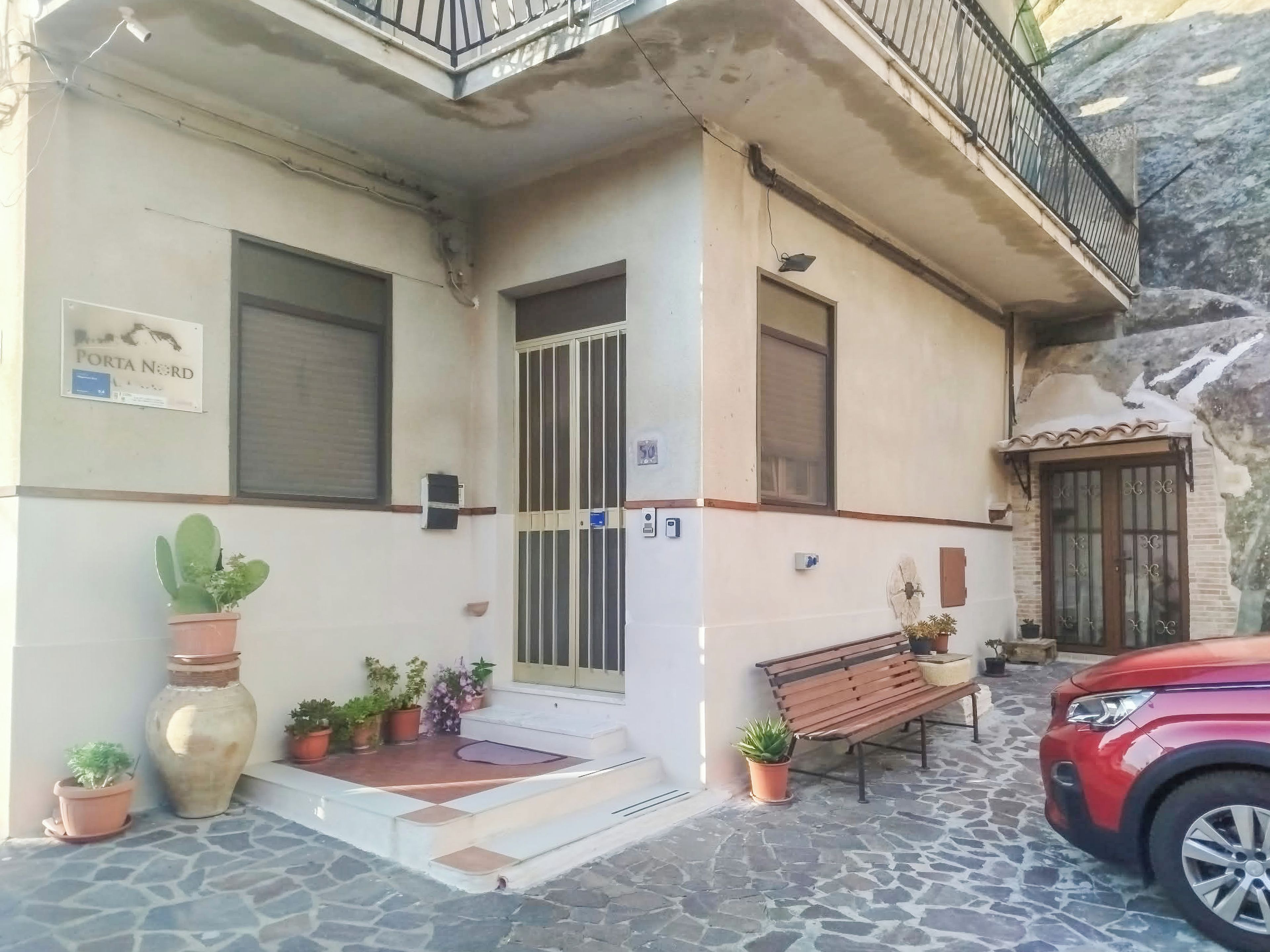 A building with a sign reading 'PORTA NORD,' a gated entrance, a bench, and a parked red car.