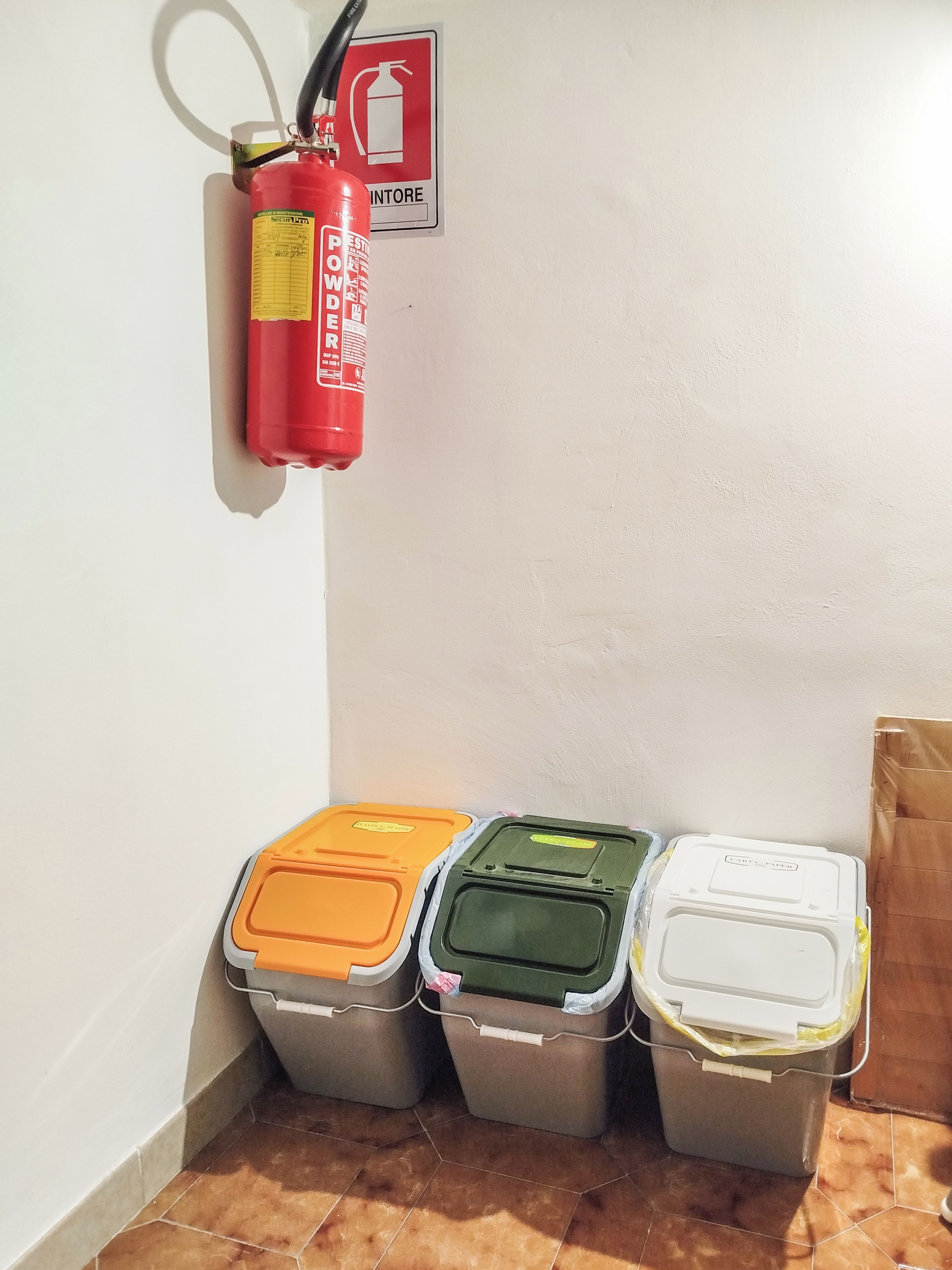 A fire extinguisher mounted on a wall with a fire safety sign, and three trash bins with different colored lids.