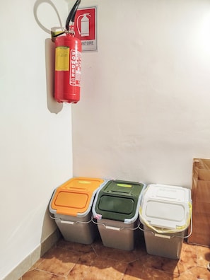 Un extintor colgado en la pared con un cartel de seguridad contra incendios, y tres contenedores de basura con tapas de diferentes colores.