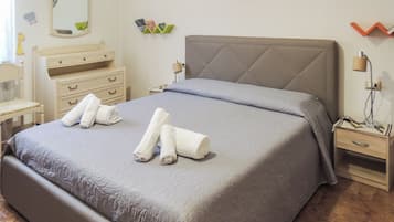 Ein ordentlich bezogenes Bett mit zwei aufgerollten Handtüchern, ein Nachttisch mit Lampe, eine Wanduhr und eine kleine Ablage mit Büchern.