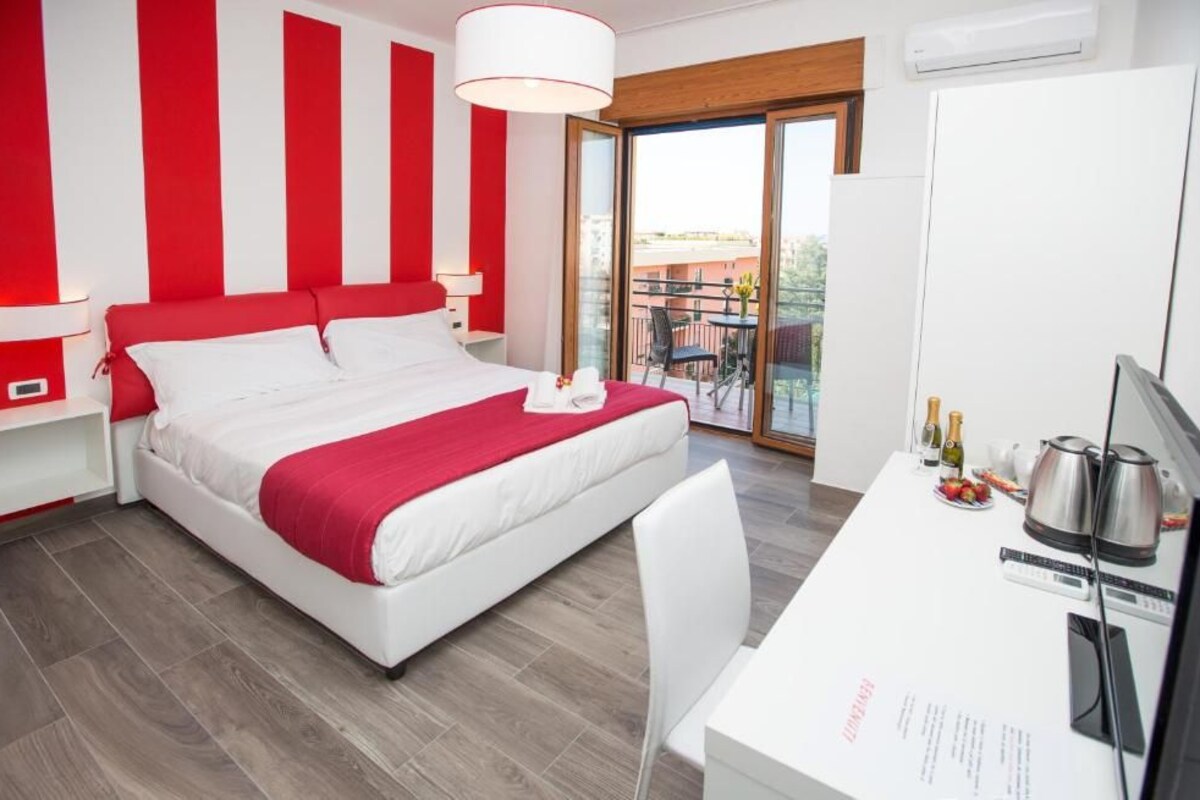 A hotel room with a bed, a desk with a computer, a chair, a table with food and drinks, and a balcony with a view.