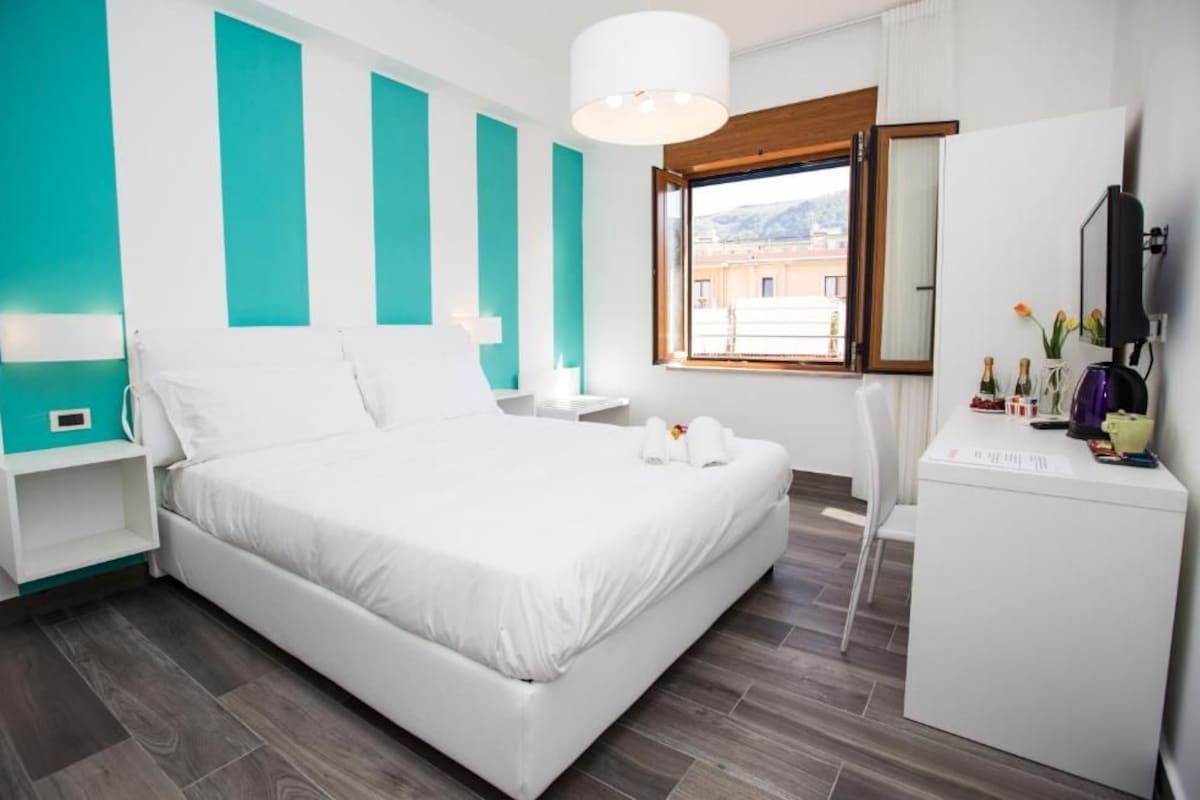 A modern hotel room with a large bed, a desk with a mirror, and a window with a view of buildings.