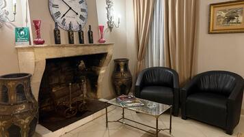 A room with a fireplace, two black leather chairs, a table with a magazine, and a large wall clock.