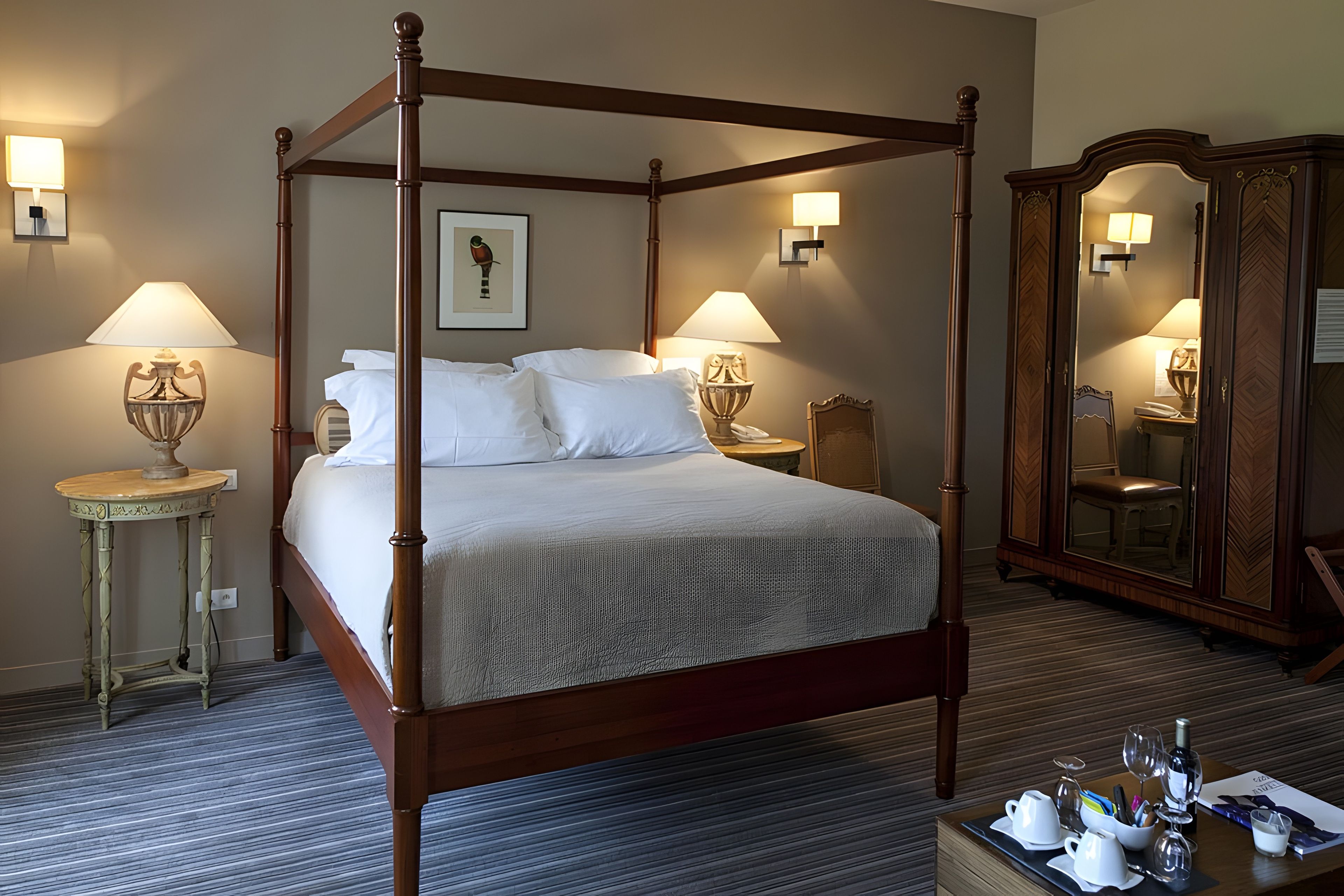 A four-poster bed with white linens, two bedside lamps, a framed picture on the wall, and a wooden wardrobe with a mirror.
