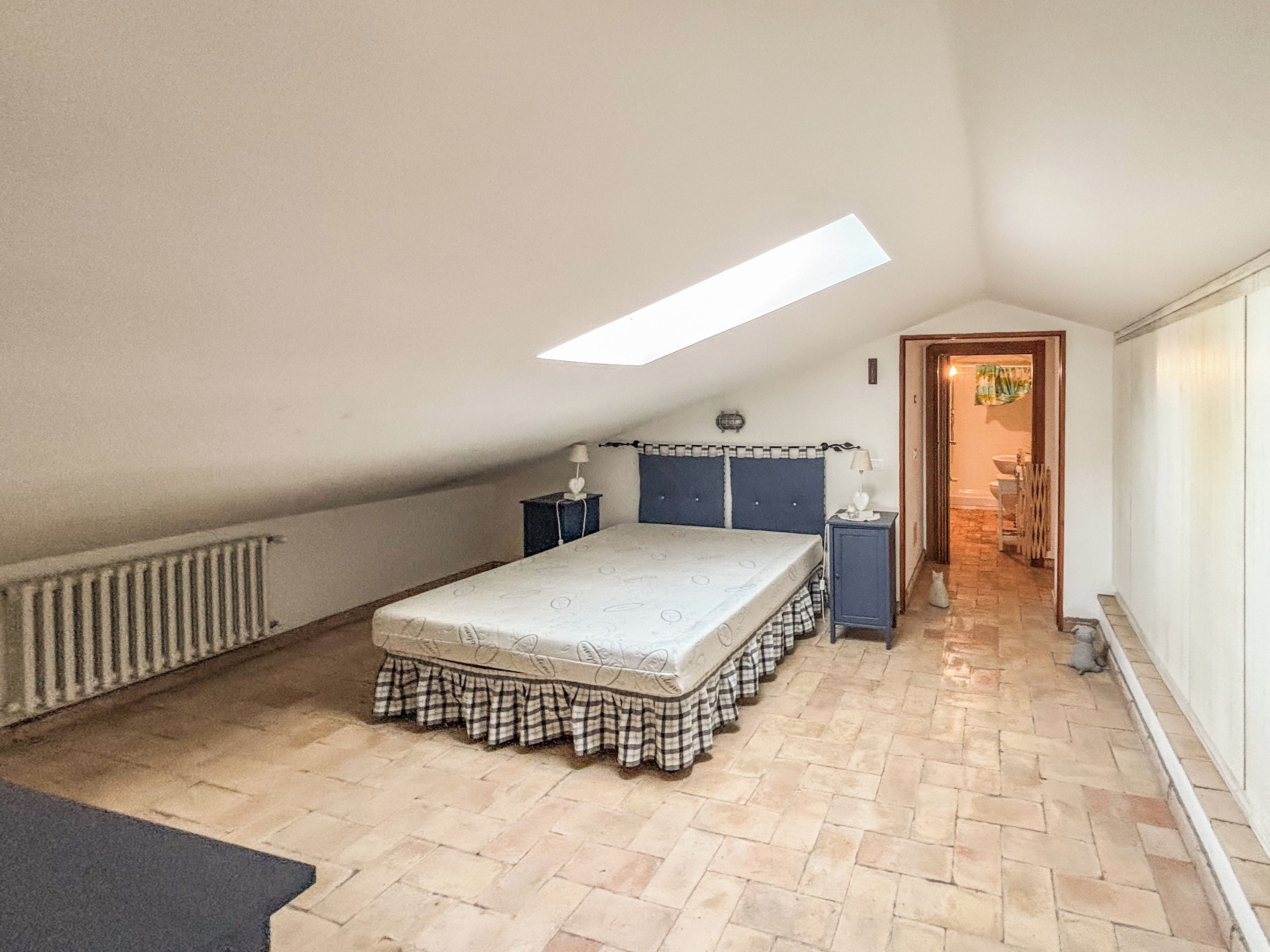 Ein Schlafzimmer mit schrägen Wänden, einem Bett mit kariertem Bettlaken, einem Nachttisch, einer Lampe und einer Heizung.