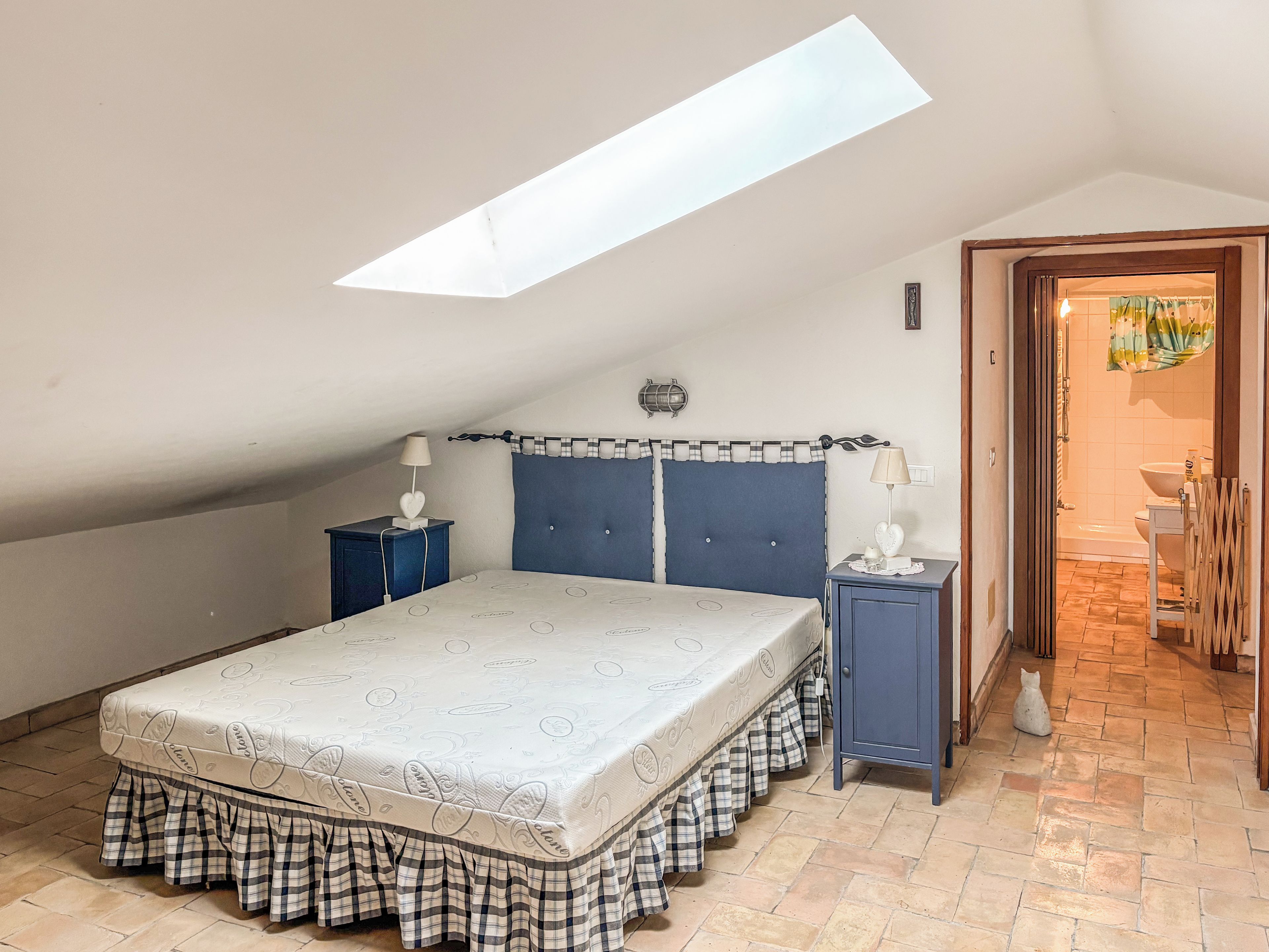 Un dormitorio con techo inclinado, una cama con falda escocesa, dos mesitas de noche con lámparas y vistas al baño por una puerta abierta.