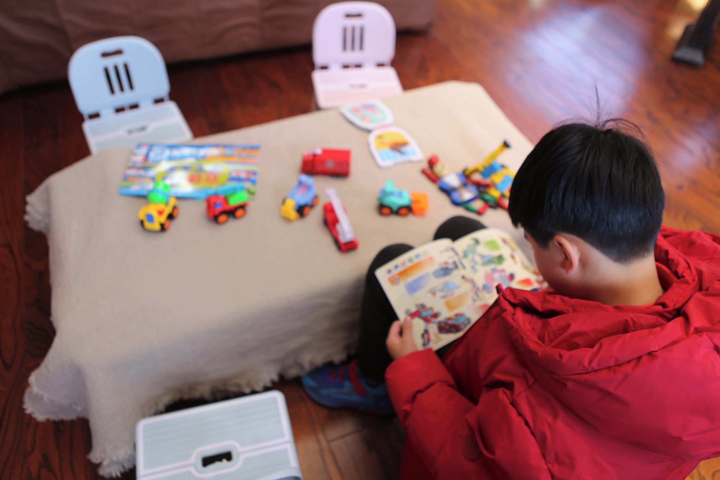 A child sitting on a carpeted floor, reading a book with toys and a blanket around.
