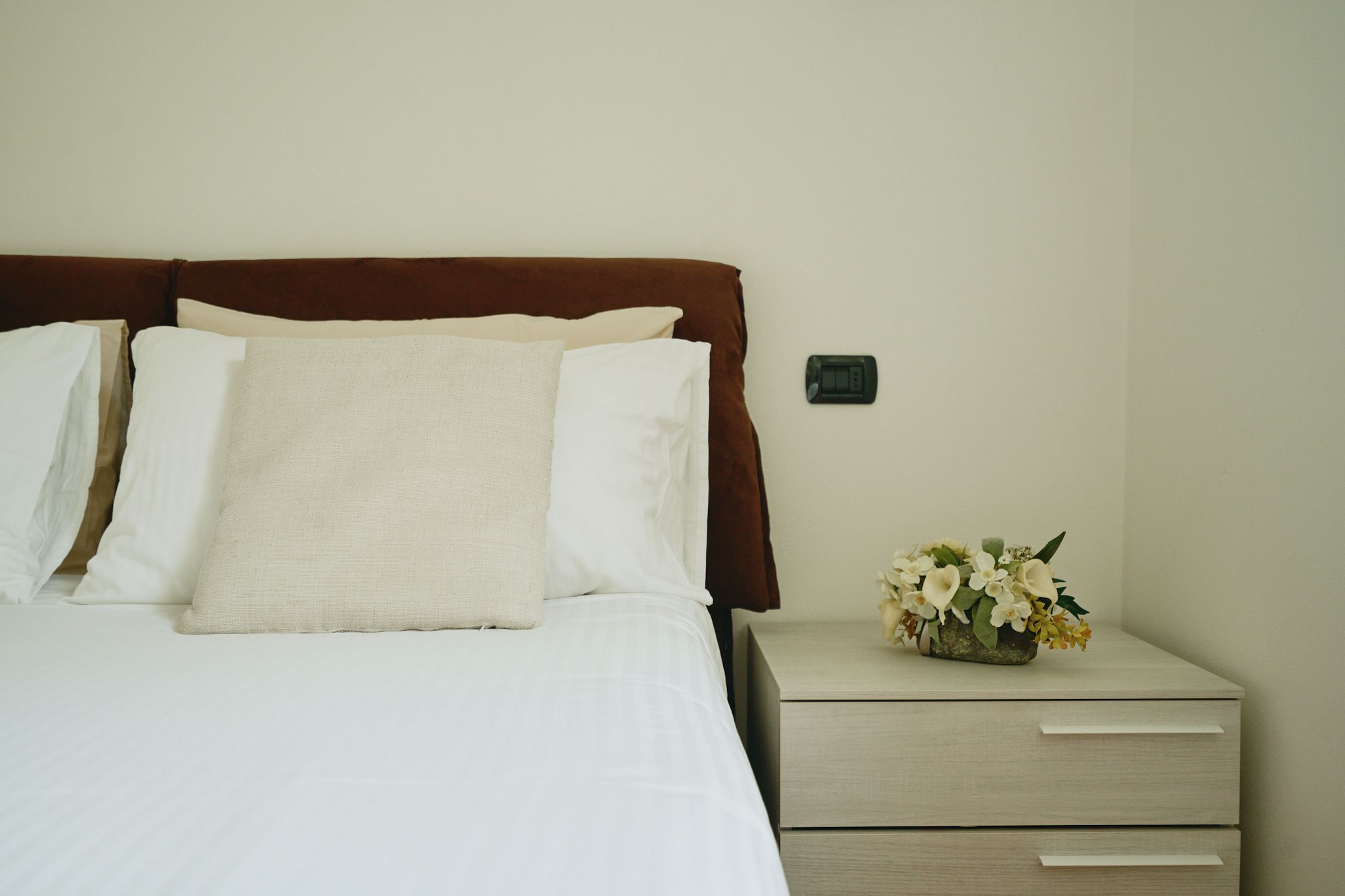 Un lit bien fait, avec du linge de lit blanc et une tête de lit marron, une petite table de chevet ornée d’un vase de fleurs et une prise électrique fixée au mur.