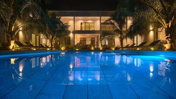 Vista nocturna de un hotel con piscina, palmeras y un edificio de dos pisos con balcones.