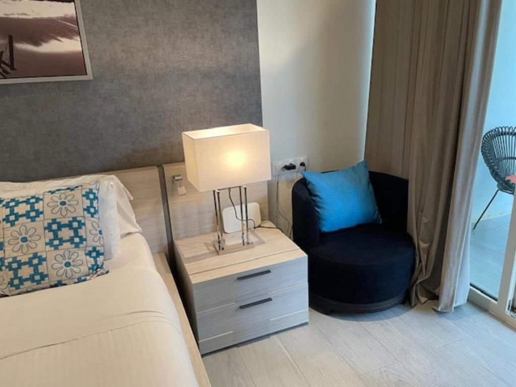 Una habitación de hotel con cama, mesita de noche con lámpara, una silla y un ventanal con cortinas.