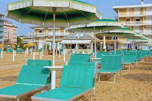 A beach with rows of green lounge chairs and umbrellas, a sandy beach, and a building with balconies in the background. - Hotel Jalisco (Lido di Jesolo)