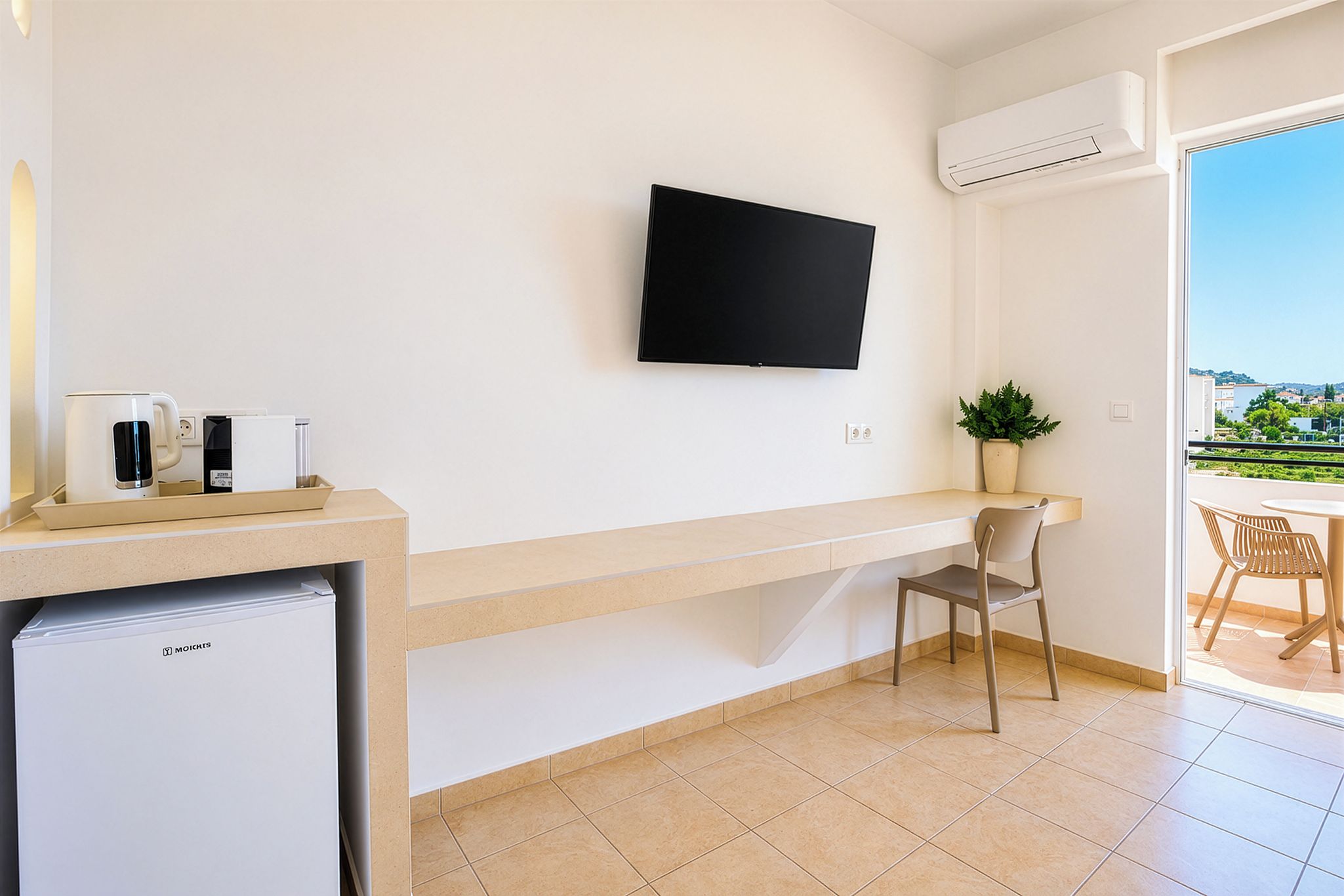A modern room with a flat-screen TV mounted on the wall, a small kitchenette with a refrigerator, a chair, and a potted plant.