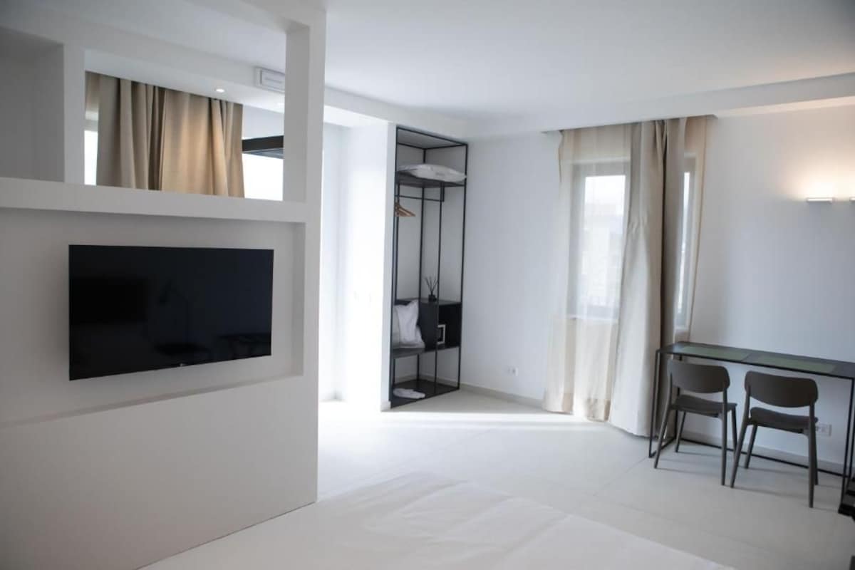 A modern hotel room with a flat-screen TV mounted on the wall, a desk with two chairs, and a walk-in closet with a mirror.