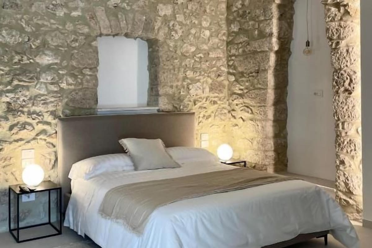 A bedroom with a stone wall, a bed with white bedding, two bedside lamps, and a nightstand.