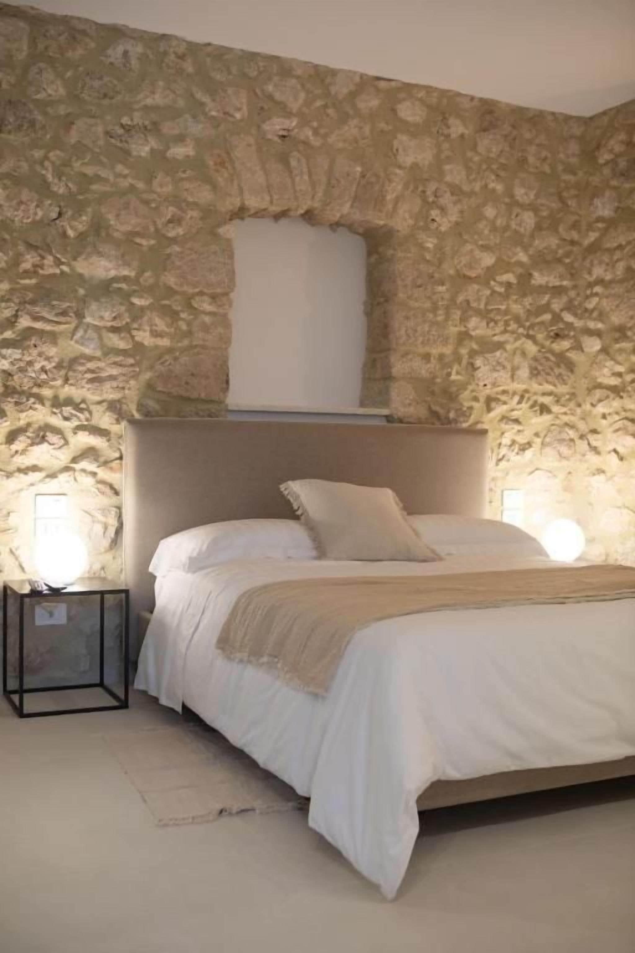 Una camera da letto con parete in pietra, un letto con biancheria bianca e un comodino con una lampada.