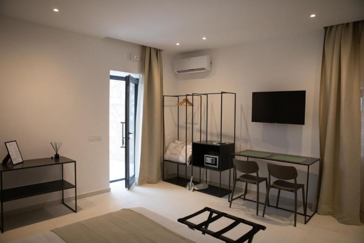 A modern hotel room with a bed, a desk, two chairs, a TV, and an air conditioning unit.