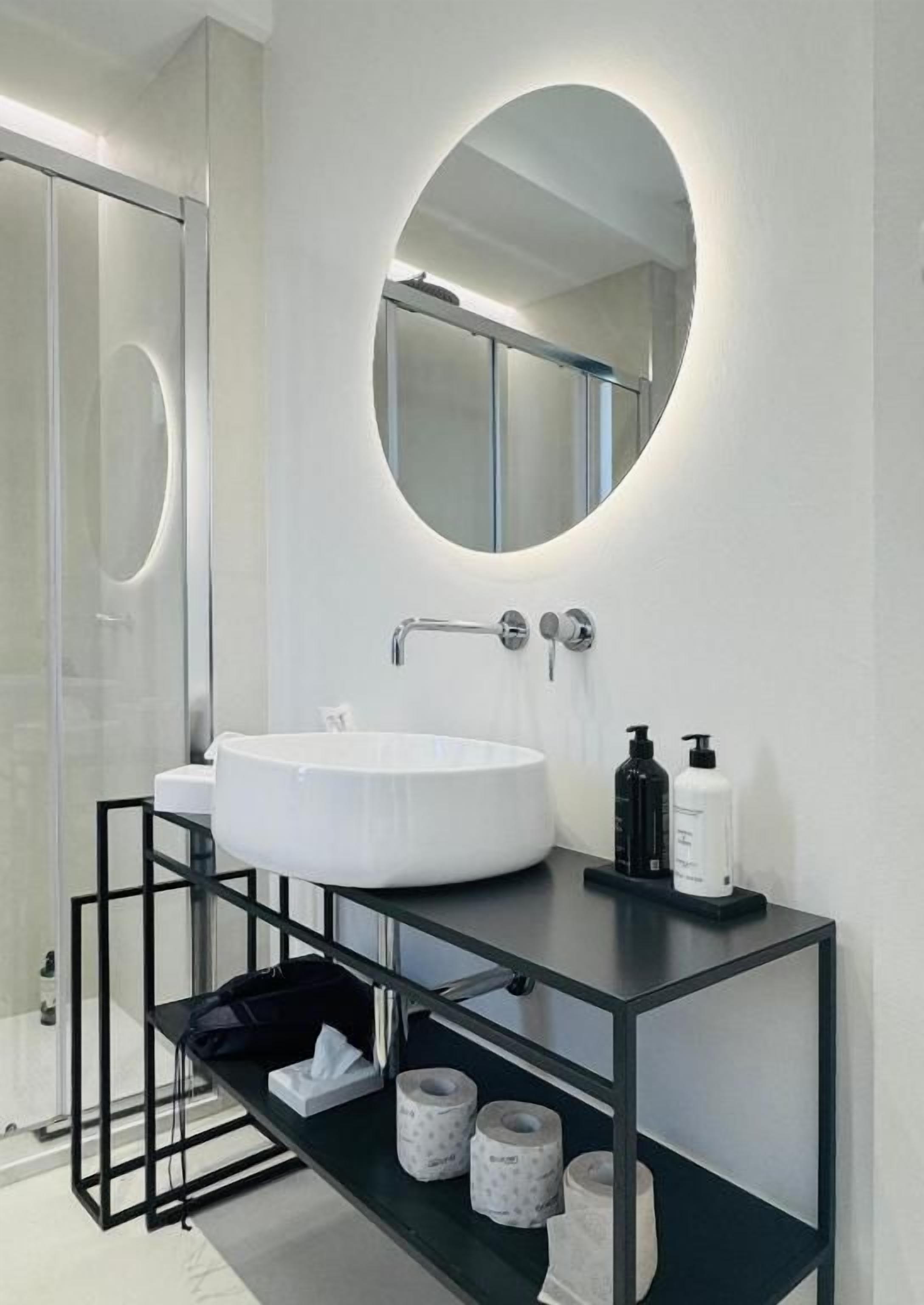 A modern bathroom with a round mirror, a white sink, and a black shelf with toiletries.