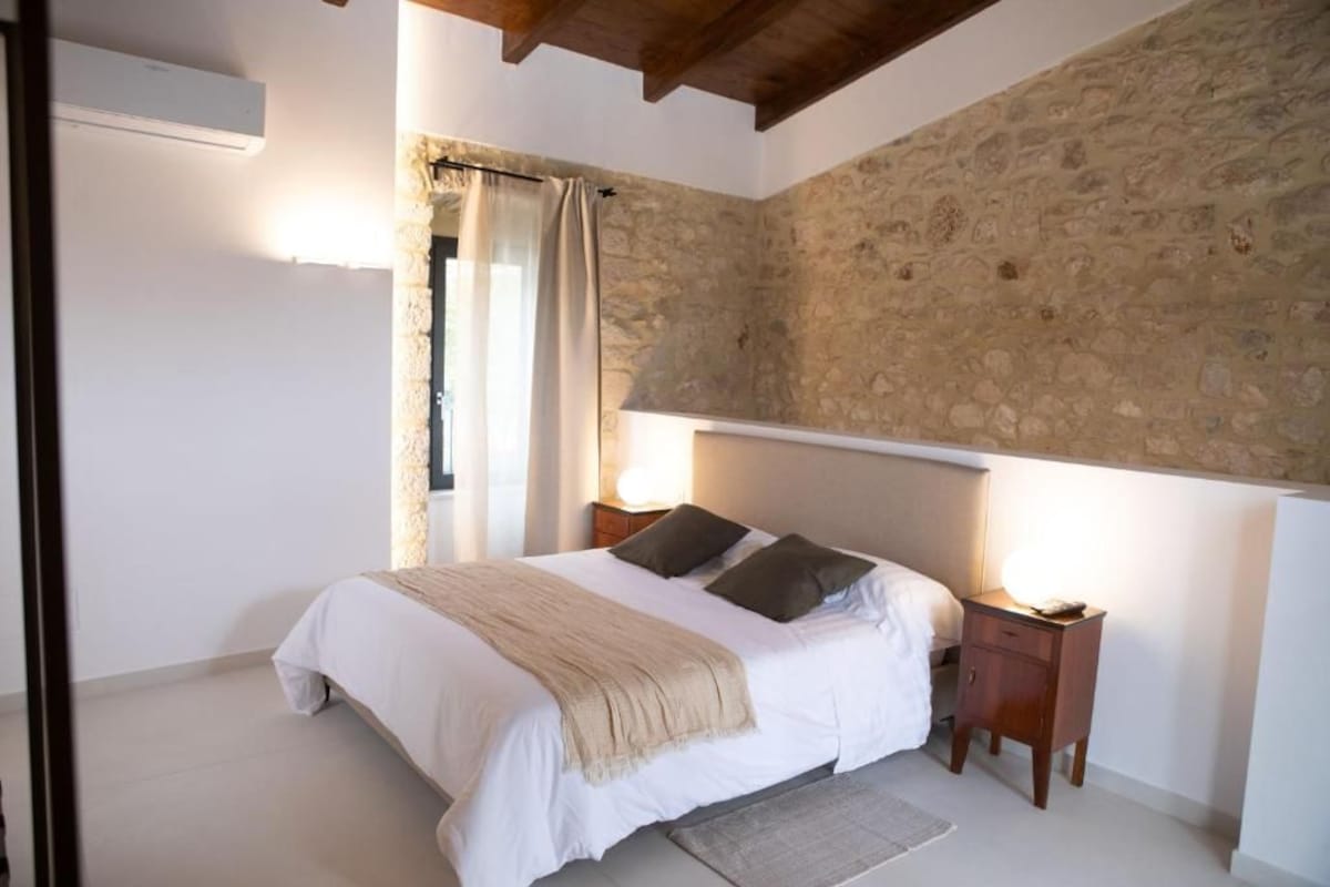 A bedroom with a stone wall, a bed with a white bedspread, two pillows, a bedside table with a lamp, and a window with curtains.