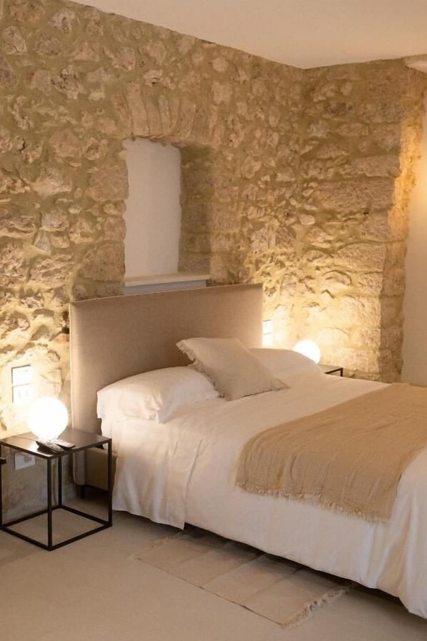 A bedroom with a stone wall, a bed with white bedding, a bedside table with a lamp, and a headboard.