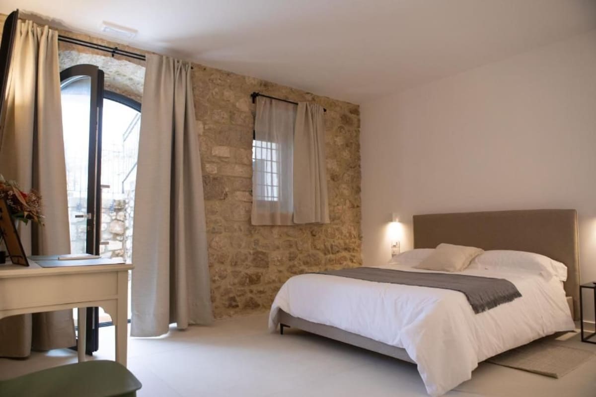 A bedroom with a stone wall, a bed with white bedding, a desk with a chair, and a window with curtains.