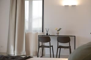 A room with a bed, a desk with two chairs, a window with curtains, and a wall-mounted light fixture.