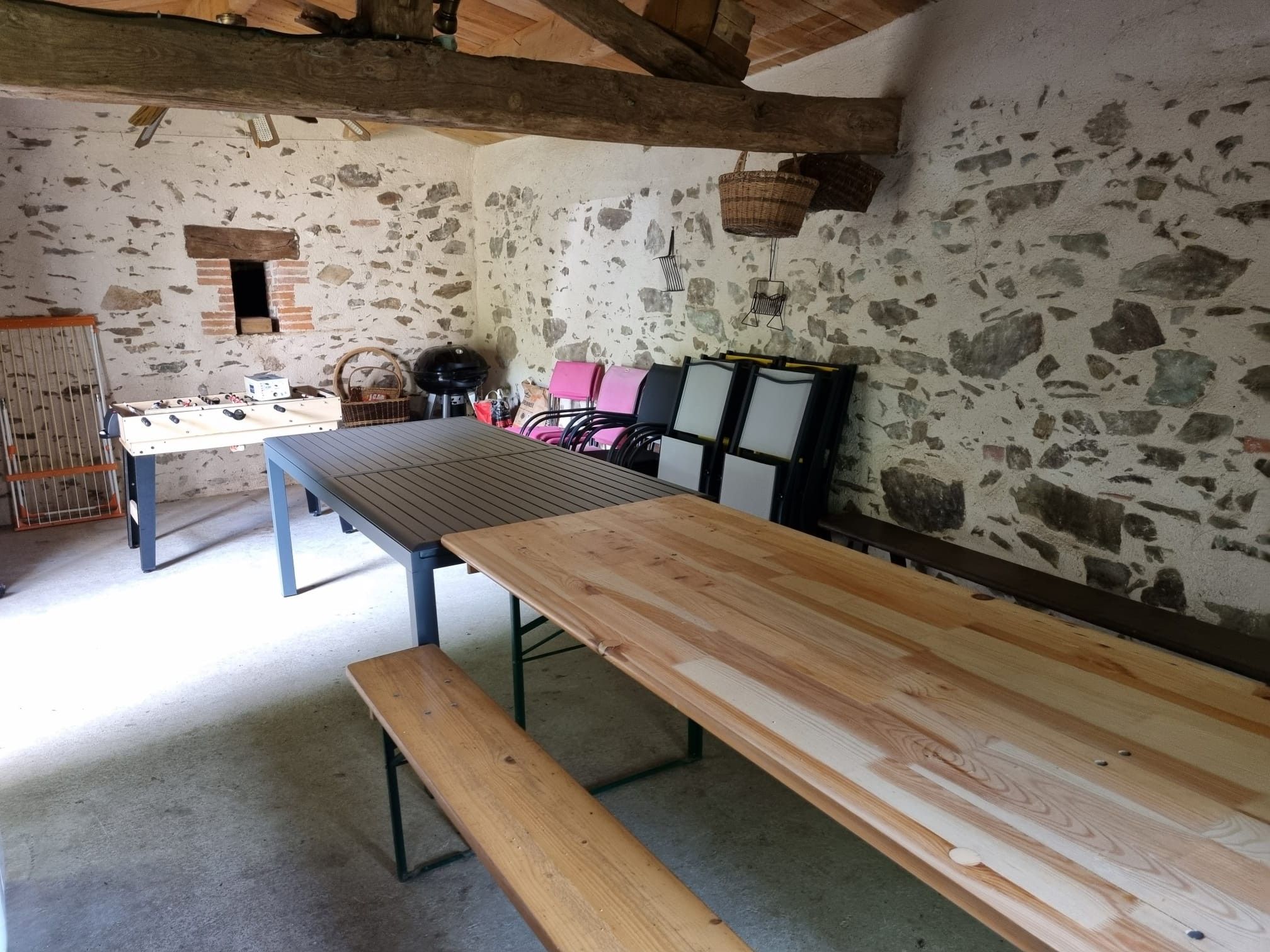 Ein rustikaler Essbereich mit einem langen Holztisch und Bänken, einem Kicker und einem Backofen im Hintergrund.