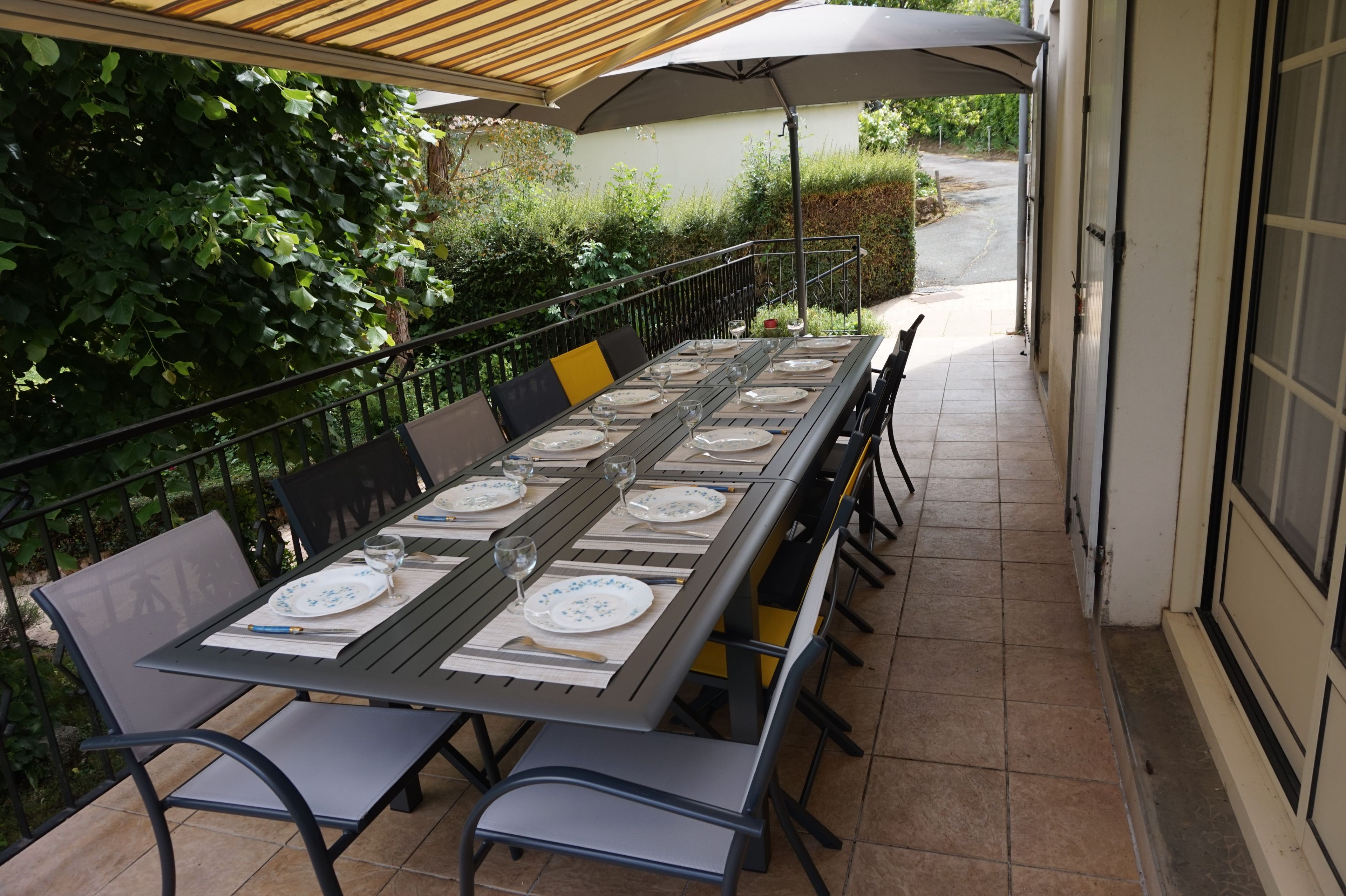 Ein Essbereich im Freien mit einem langen Tisch, gedeckt für sechs Personen, umgeben von Stühlen unter einem beschatteten Sonnenschirm.