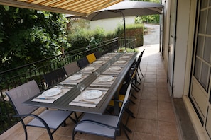 Ein Essbereich im Freien mit einem langen Tisch, gedeckt für sechs Personen, umgeben von Stühlen unter einem beschatteten Sonnenschirm.