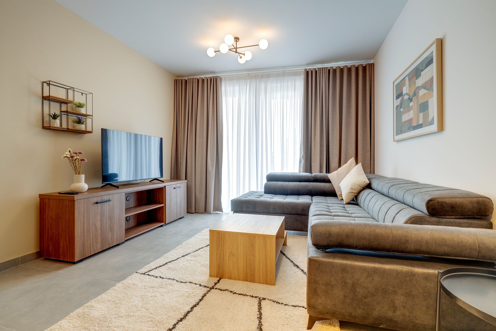 Una sala de estar moderna con un sofá seccional, un televisor sobre un mueble de madera y un amplio ventanal con cortinas.