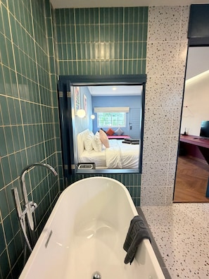 Ein modernes Badezimmer mit einer großen Badewanne, einer Dusche und einem Spiegel, der ein Schlafzimmer mit Bett und Fenster reflektiert.
